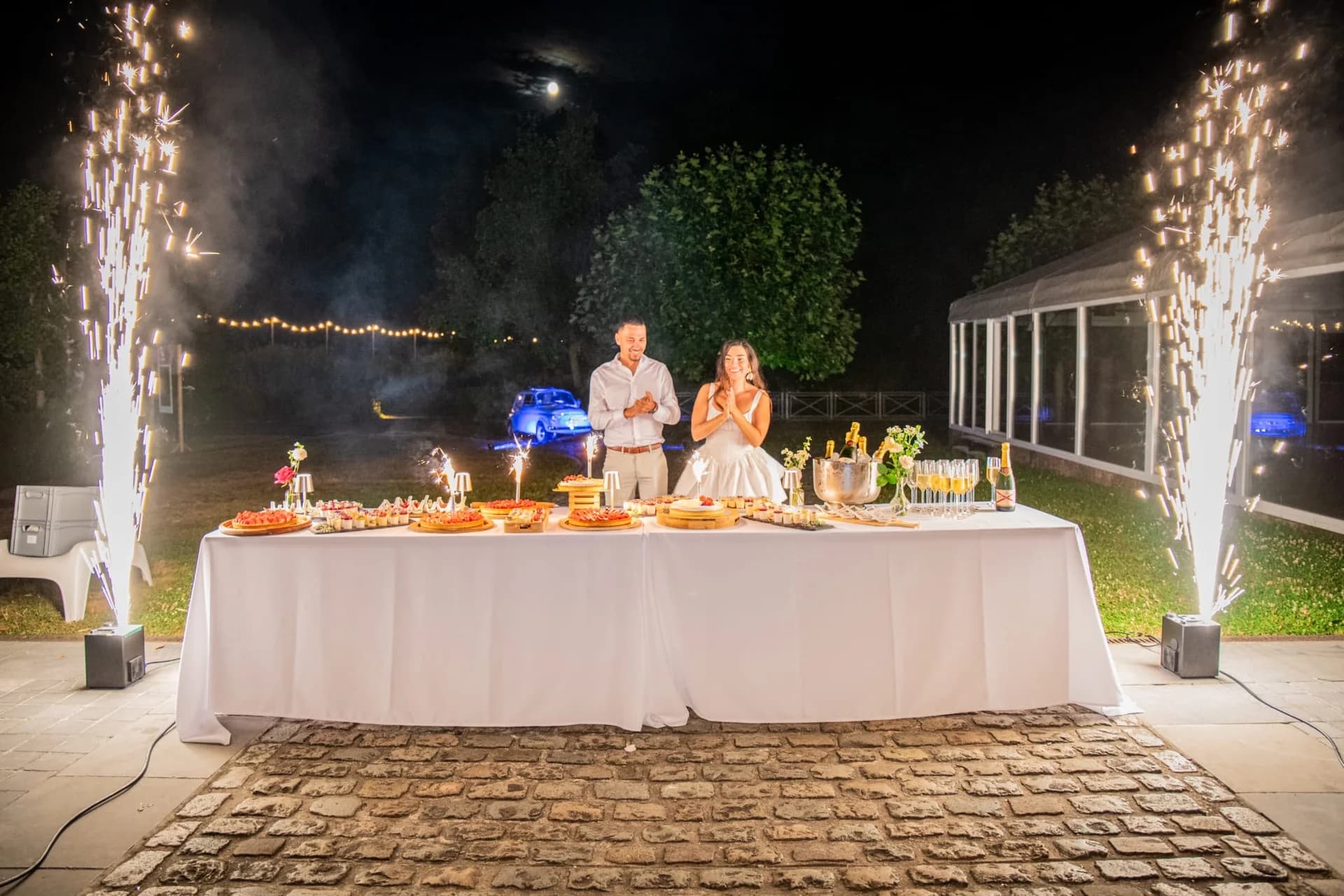 Les mariés devant leur buffet de desserts, entourés de deux gerbes d'étincelles en extérieur