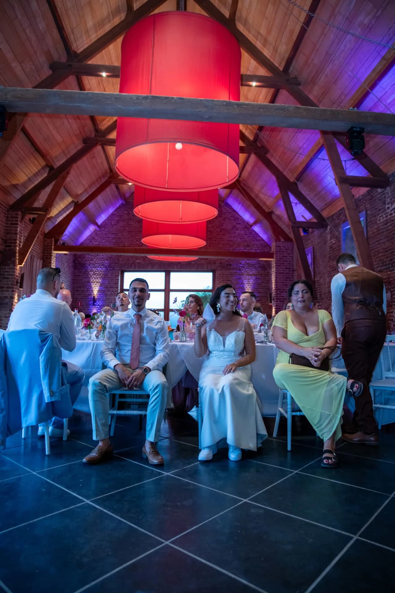 Les mariés et leurs invités dans une salle aux poutres et aux murs éclairés de violet